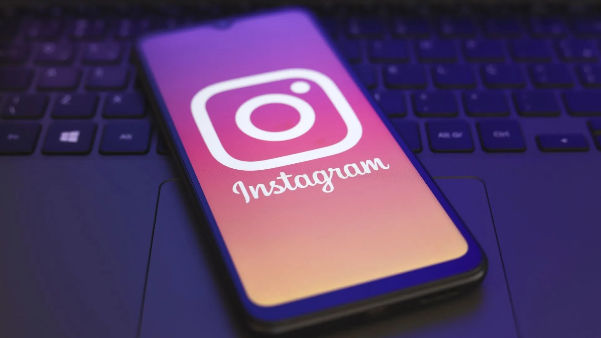 یک گوشی موبایل روی صفحه‌کلید لپ‌تاپ قرار دارد و صفحه‌ی گوشی لوگو و نام «Instagram» را با زمینه‌ی گرادیانی صورتی تا نارنجی نمایش می‌دهد.