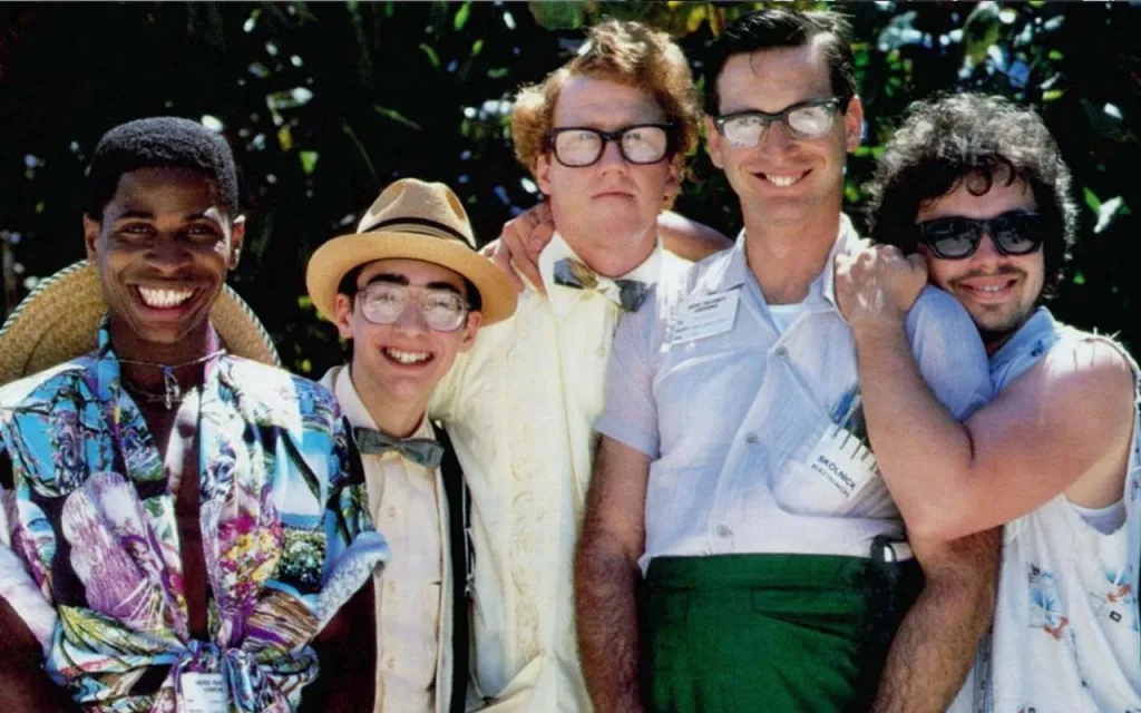  تصویری از فیلم Revenge Of The Nerds 1984  که در ان  افراد با لباس‌های رنگارنگ و سبک گرمسیری کنار هم ایستاده‌اند، برخی از آن‌ها کلاه حصیری یا لباس‌های طرح‌دار به تن دارند. پس‌زمینه تصویر شامل شاخ و برگ‌های سبز و روشن است که حال و هوای فضای طبیعی و گرمسیری را نشان می‌دهد.





