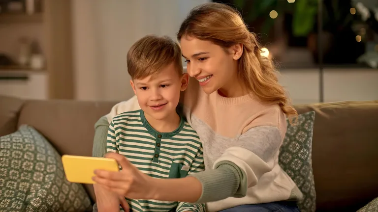 یک زن و کودک روی کاناپه نشسته‌اند و با یک گوشی زرد مشغول گرفتن عکس سلفی هستند. در پس‌زمینه، فضای خانه با نور گرم و گیاهان تزئینی دیده می‌شود که حس آرامش را منتقل می‌کند.