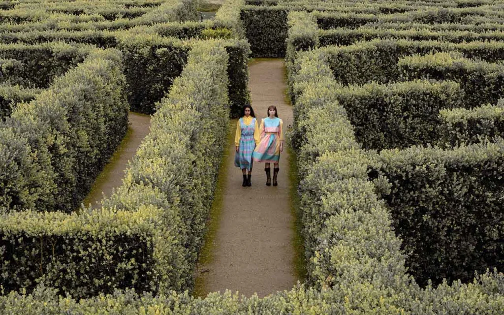 دو زن در حال قدم زدن در میان راهروهای باریک یک باغ پر از پرچینهای بلند و سبز هستند.