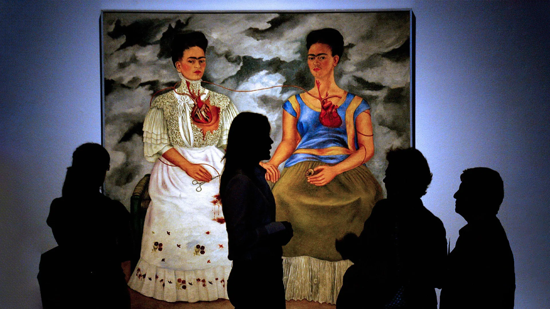 تابلویی از نقاشی مشهور «دو فریدا» اثر فریدا کالو به نمایش گذاشته شده که دو زن با لباس‌های متفاوت کنار هم نشسته‌اند و قلب‌های آنها از طریق رگ‌های باز به هم وصل شده است. فضای پس‌زمینه ابری و تیره است و چند نفر در مقابل نقاشی ایستاده‌اند و آن را تماشا می‌کنند.