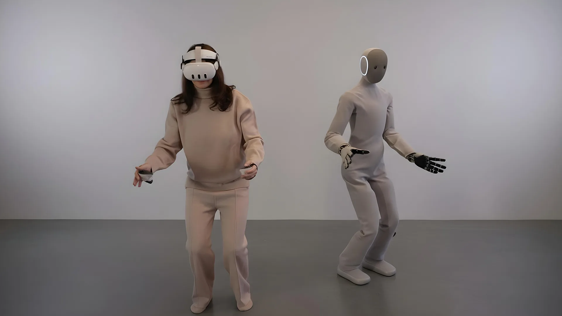 زنی با لباس کرم‌رنگ و عینک واقعیت مجازی روی چشمانش ایستاده و در حال حرکت است. کنار او رباتی انسان‌نما با بدنی خاکستری و دستان مکانیکی دیده می‌شود که حالت بدنی مشابه او دارد.