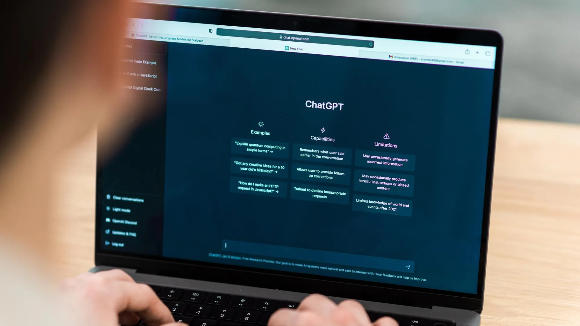 فردی پشت لپ‌تاپی نشسته و صفحه‌ی اصلی ChatGPT روی نمایشگر باز است. در صفحه، بخش‌هایی با عنوان‌های «Examples»، «Capabilities» و «Limitations» دیده می‌شود که توضیحاتی درباره توانایی‌ها و محدودیت‌های چت‌جی‌پی‌تی ارائه می‌دهد.