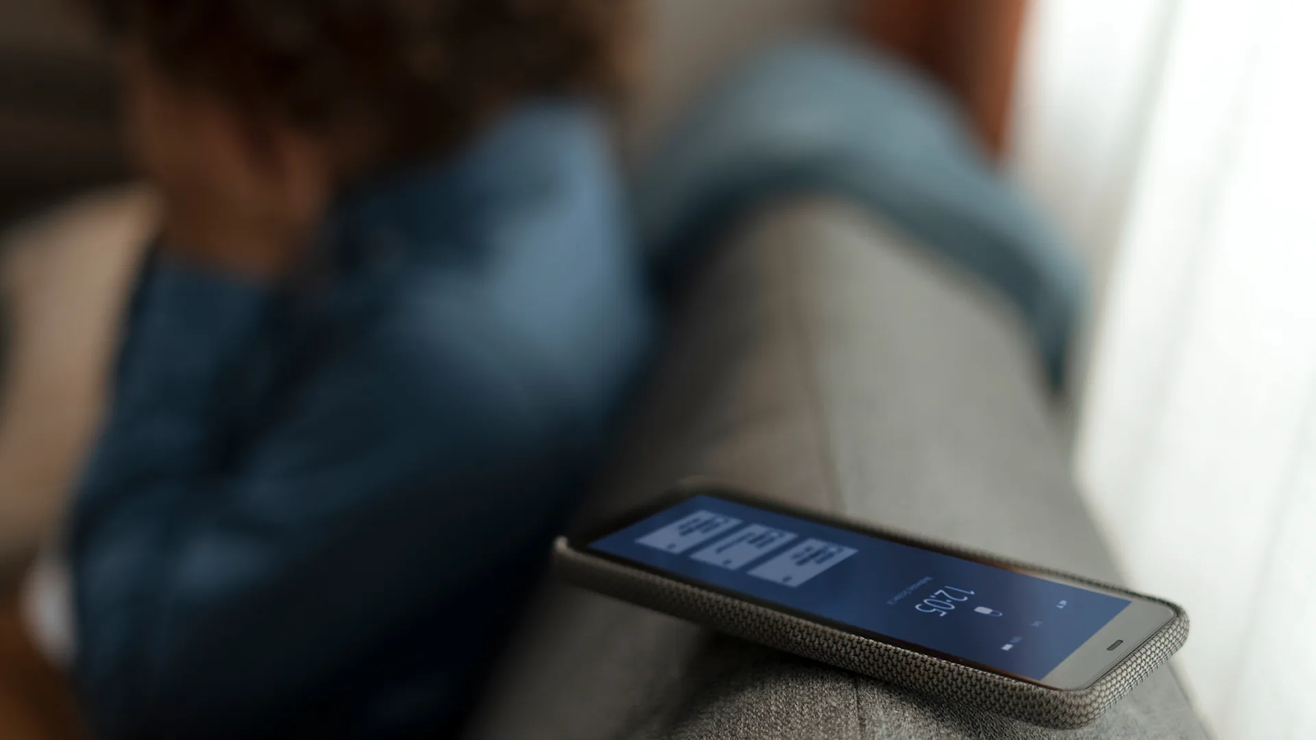 تلفن همراهی روی مبل قرار دارد که صفحهی تماس فعال روی آن دیده میشود. در پسزمینه، فردی با لباس آبی بهصورت تار و ناراحت نشسته و دستش را روی صورت خود گذاشته است.