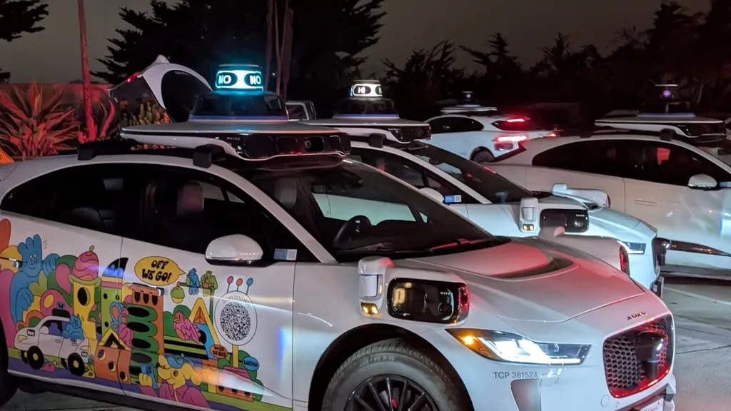  چند خودروی خودران سفیدرنگ در شب دیده می‌شوند که روی سقف آن‌ها حسگرها و نوشته‌های دیجیتالی مانند “NO” و “HI” قرار دارد. خودروی جلویی با طراحی کارتونی رنگارنگ تزئین شده و بخشی از پروژه‌ای متعلق به شرکت وایمو (Waymo)، وابسته به گوگل، است.