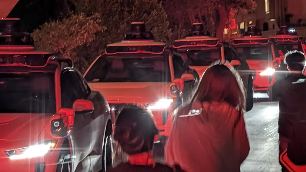 در این تصویر شبانه، بیش از ۵۰ خودروی بدون راننده با چراغ‌های روشن در خیابانی باریک صف کشیده‌اند. چند نفر از پشت سر دیده می‌شوند که به سمت این خودروها نگاه می‌کنند یا از آن‌ها عکس می‌گیرند، و نور قرمز محیط، صحنه‌ای غیرعادی و کمی نگران‌کننده ایجاد کرده است.