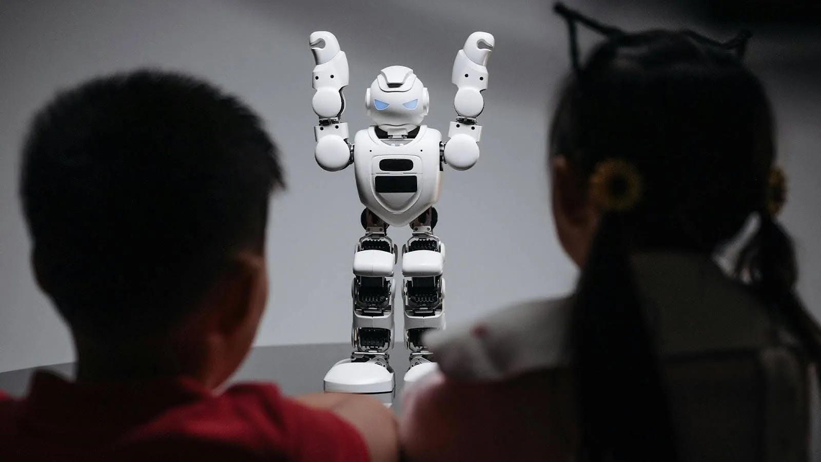یک ربات کوچک سفیدرنگ را نشان می‌دهد که دست‌های خود را بالا برده و حالت شادی دارد. دو کودک یکی دختر یکی پسر در مقابل ربات نشسته‌اند و با کنجکاوی به آن نگاه می‌کنند.