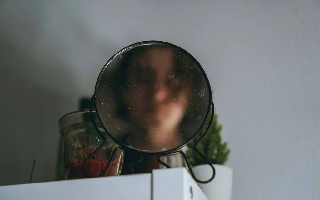 ک آینه گرد روی پایه فلزی را نشان میدهد که روی یک سطح سفید قرار گرفته است و تصویر مبهم یک دختر در اینه است. در کنار آینه، یک ظرف شیشهای با طرحهایی روی آن و یک گیاه کوچک سبزرنگ دیده میشود.