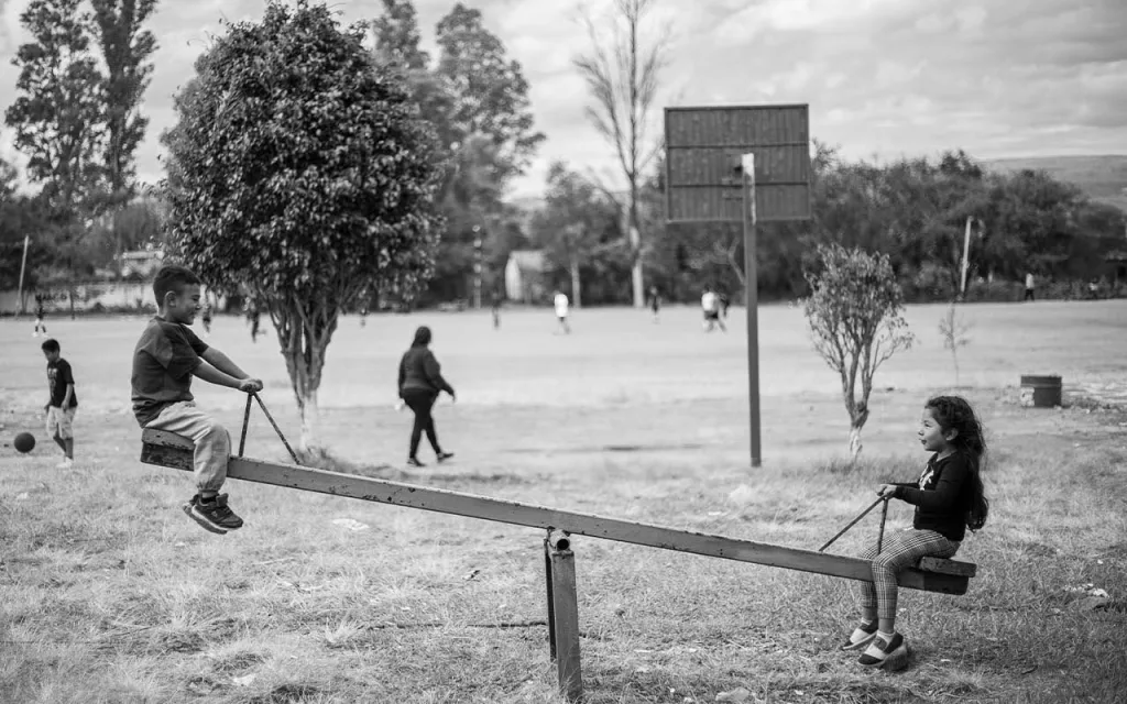 تصویر یک الاکلنگ فلزی را نشان میدهد که دو کودک در دو طرف آن نشستهاند و دختربچه در قست پایین و پسربچه در قسمت بالا قراردارند. در پسزمینه، فضای باز یک پارک دیده میشود که شامل زمین چمن، درختان و افرادی است که در حال قدم زدن یا بازی کردن هستند.