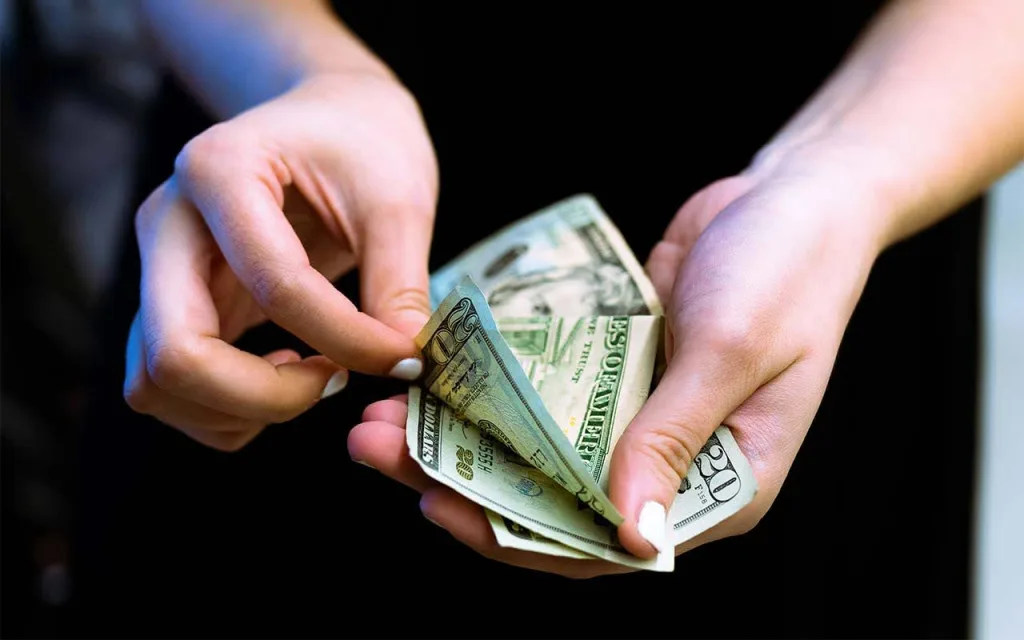 دو دست دیده میشود که تعدادی اسکناس دلار را نگه داشتهاند و یکی از اسکناسها در حال جدا شدن است. زمینه تصویر تیره است و تمرکز اصلی روی دستها و اسکناسها قرار دارد.