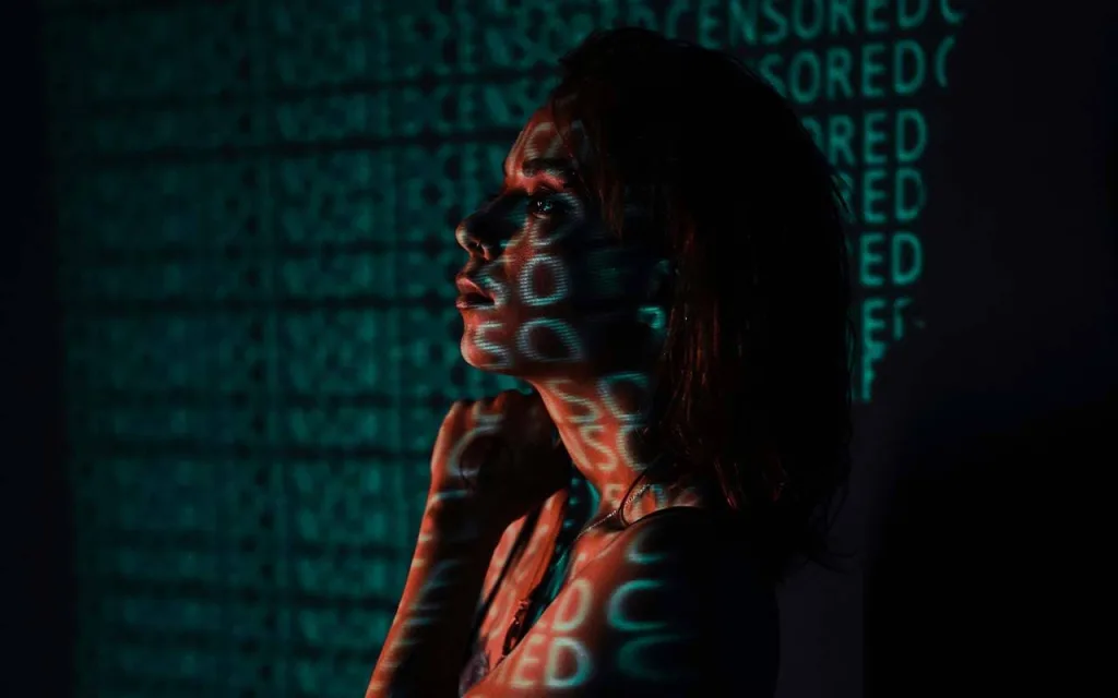 یک زن در جلو تصویر قرار دارد و کلمات “CENSORED” به رنگ سبز به صورت پراکنده روی دیوار و بدن وی دیده میشوند. نورپردازی تاریک و سایهها حس رمزآلود و مفهومی به تصویر داده است.