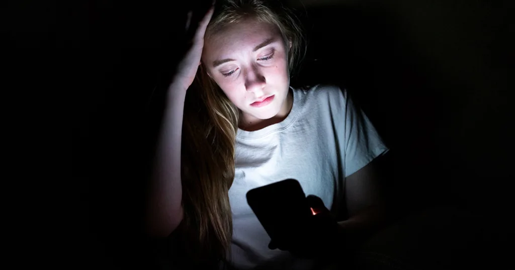 یک دختر نوجوان با لباس سفید در فضایی تاریک نشسته و نور ضعیفی از صفحه گوشی همراهش به اطراف تابیده شده است. دست او روی سرش قرار دارد و حالتی از خستگی یا تفکر را نشان می‌دهد.