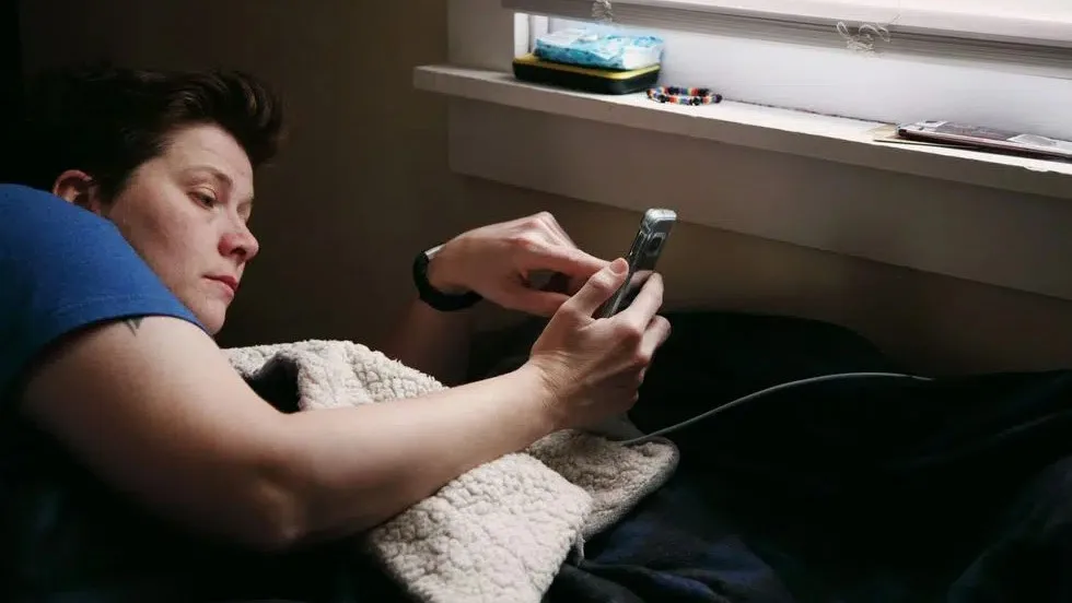 فردی روی تخت دراز کشیده و یک تلفن همراه را با هر دو دست نگه داشته است. کنار پنجره‌ای در پس‌زمینه، چند وسیله کوچک مانند جعبه دستمال کاغذی و مهره‌های رنگی دیده می‌شود.