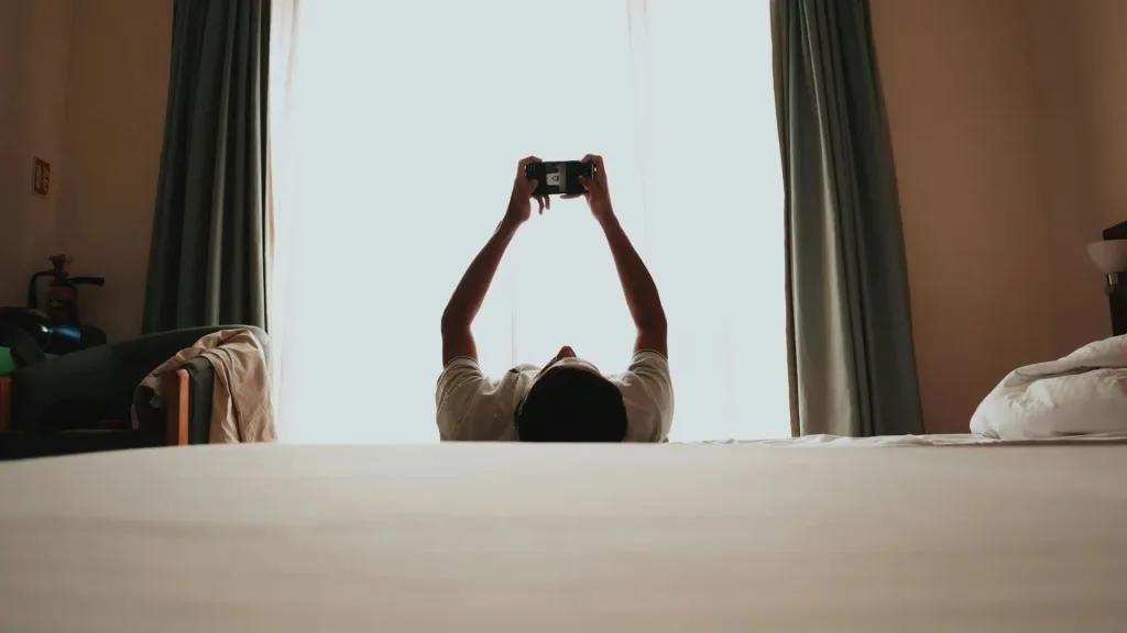 فردی به پشت روی تخت دراز کشیده و دستانش را بالا برده تا با گوشی موبایل کار کند.