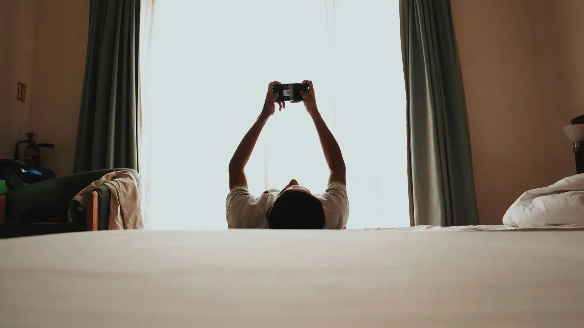 فردی به پشت روی تخت دراز کشیده و دستانش را بالا برده تا با گوشی موبایل کار کند.