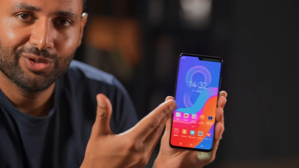 فردی گوشی موبایلی را نشان می‌دهد که صفحه آن یک رابط کاربری رنگارنگ با ساعت 14:32 و چندین آیکون برنامه‌ در پایین صفحه نمایش داده شده است.