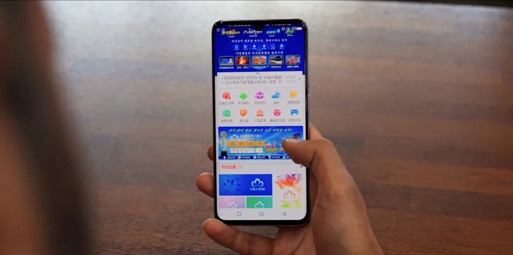تصویر یک گوشی هوشمند را نشان می‌دهد که در دست فردی قرار دارد و صفحه آن یک اپلیکیشن یا پورتال با رابط کاربری کاملاً کره‌ای و رنگی را نمایش می‌دهد. آیکون‌های متعدد در صفحه دیده می‌شود که شبیه نسخه داخلی و محدودشده‌ای از فروشگاه یا شبکه خدمات آنلاین است.