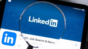 لوگوی لینکدین با متن سفید “LinkedIn” روی پس‌زمینه آبی دیده می‌شود که طرح‌هایی از خطوط و نقاط شبکه‌ای را در خود دارد. یک ذره‌بین بخش میانی تصویر را بزرگ کرده و بر نام لینکدین تأکید می‌کند.