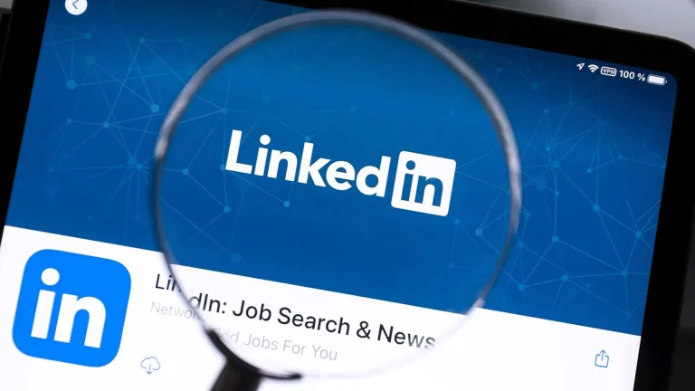 لوگوی لینکدین با متن سفید “LinkedIn” روی پس‌زمینه آبی دیده می‌شود که طرح‌هایی از خطوط و نقاط شبکه‌ای را در خود دارد. یک ذره‌بین بخش میانی تصویر را بزرگ کرده و بر نام لینکدین تأکید می‌کند.