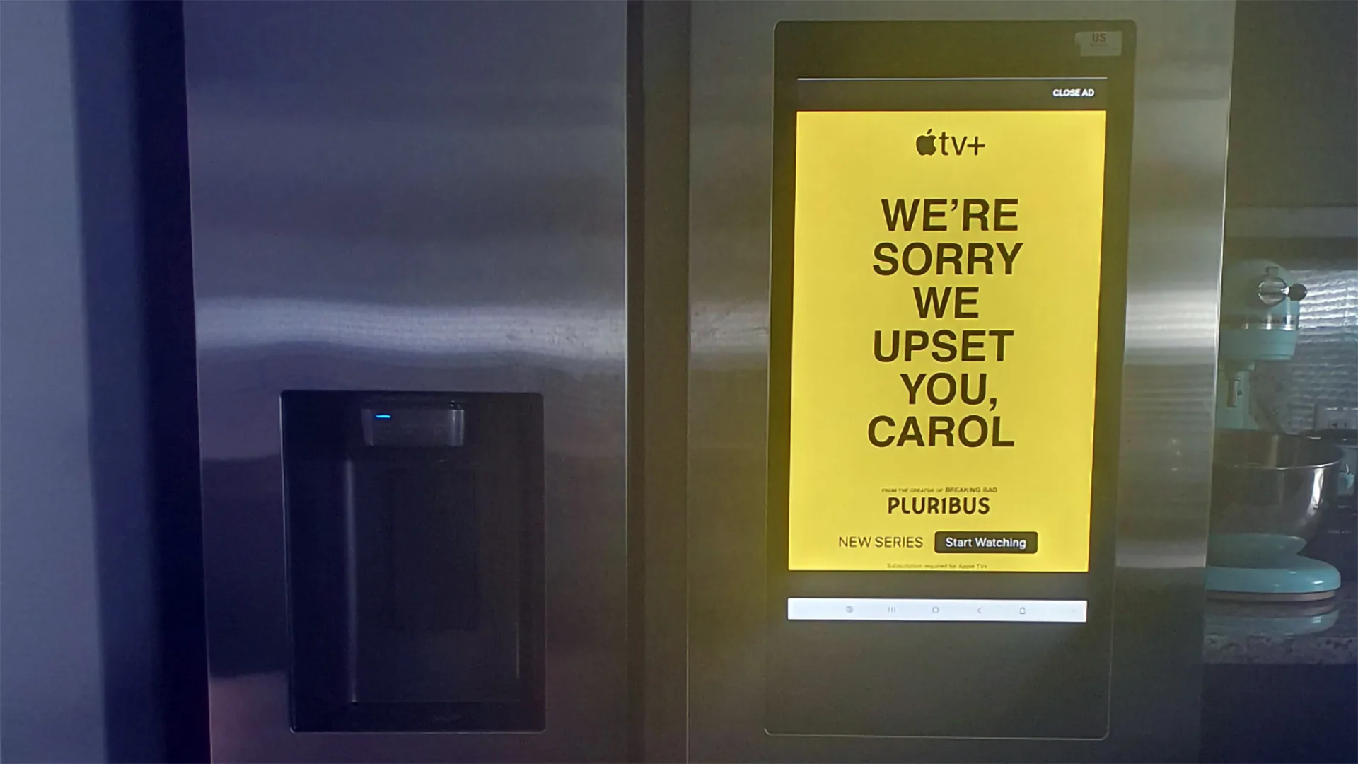 تصویری از یک یخچال هوشمند دیده می‌شود که روی صفحه نمایش آن تبلیغی با پس‌زمینه زرد و متن سیاه نمایش داده شده است. متن تبلیغ از طرف Apple TV+ است و نوشته: «کارول، بابت ناراحت کردنت معذرت می‌خواهیم»، که به معرفی یک سریال جدید به نام “Pluribus” اشاره دارد.