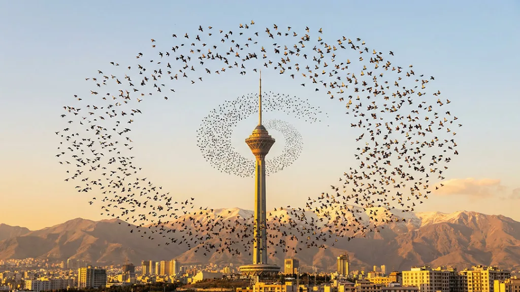 برج میلاد تهران را در پس‌زمینه‌ای از کوه‌های برفی و آسمانی روشن نشان می‌دهد، در حالی که گروهی از پرندگان به شکلی دایره‌وار و منظم در اطراف برج پرواز می‌کنند.
پرواز پرندگان حالتی غیرطبیعی و بیش از حد هماهنگ دارد که نشان می‌دهد این تصویر ممکن است با هوش مصنوعی ساخته شده باشد.