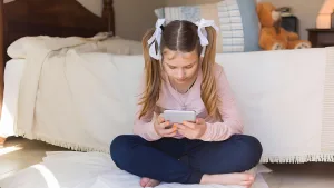 دختربچه‌ای روی زمین نشسته و با تمرکز به گوشی هوشمندش نگاه می‌کند؛ تصویری که نمادی از جذب شدن کودکان به ویدیوهای کوتاه و اسکرول بی‌نهایت است. این صحنه نشان می‌دهد چگونه استفاده مداوم از موبایل می‌تواند بر مغز کودکان، توجه و عادات روزمره آن‌ها تأثیر بگذارد.