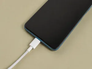 کابل سفید USB-C در حال اتصال به درگاه پایین یک گوشی هوشمند آبی‌رنگ روی سطح کرم‌رنگ