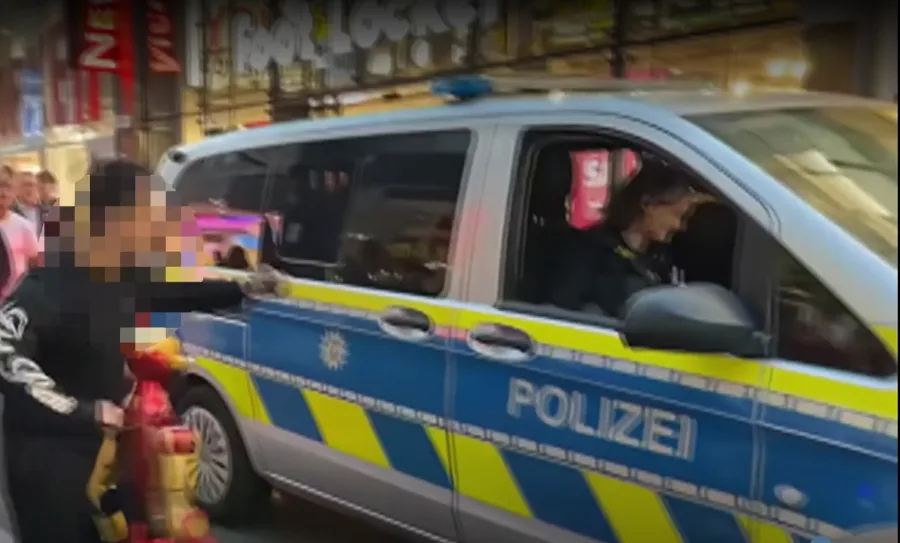 خودروی پلیس آلمان با نوشته POLIZEI در خیابان و فردی با چهره شطرنجیشده کنار ربات قرمز و طلایی